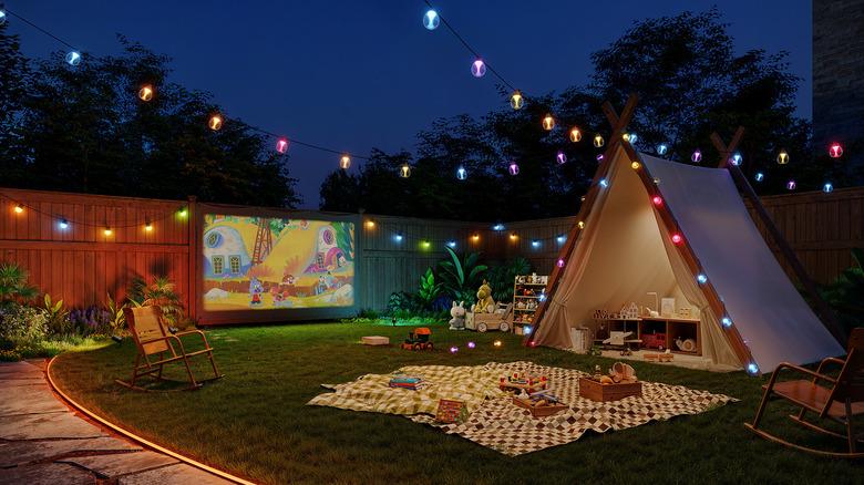 backyard at night set up with a projector movie screen, tent, blanket, and colorful Govee string lights for a happy, playful mood
