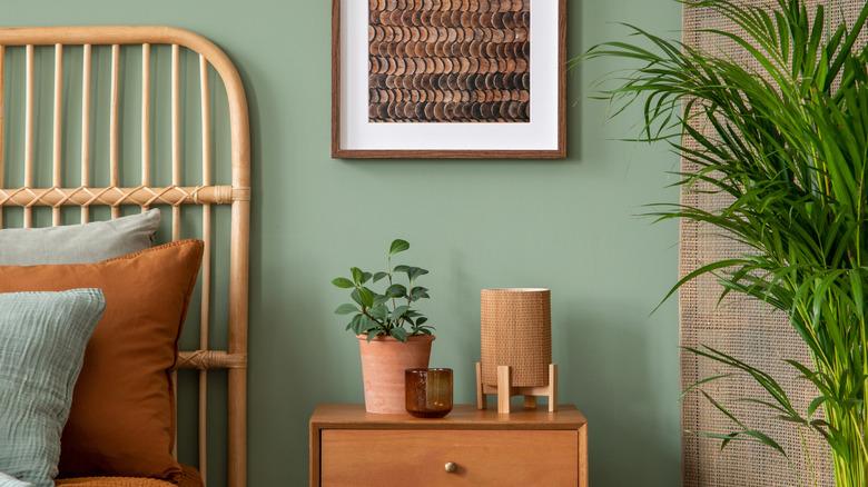 bedroom with sage green walls, a natural rattan headboard, a palm floor plant, and a wood mid-century nightstand with an artificial plant in a terracotta pot