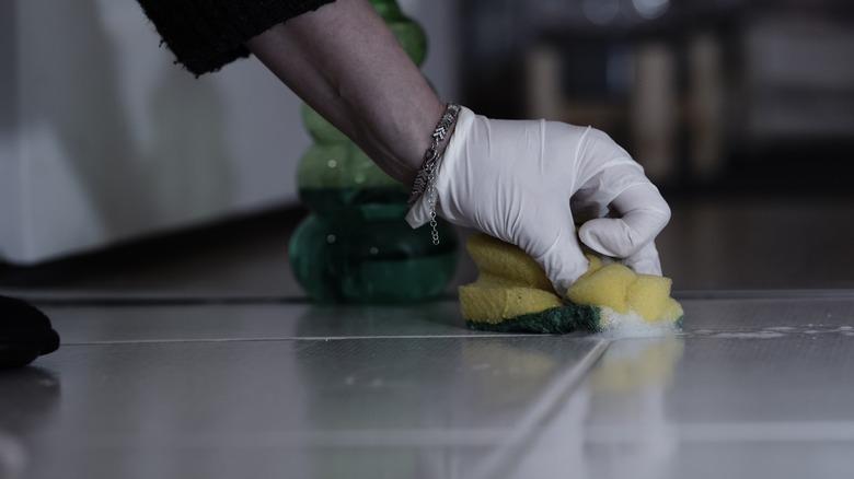A gloved hand scrubbing grout between floor tiles