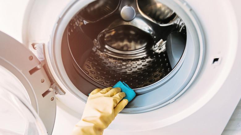 A yellow-gloved hand is wiping the rubber gasket around the opening of a front-loaded washing machine.