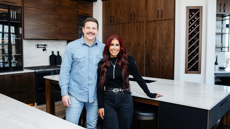 Cole and Chelsea DeBoer smiling in front of a newly remodeled kitchen with dark wood cabinets and light stone countertops