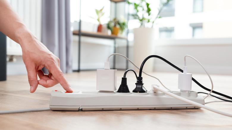 A hand turning on a white extension cord on a wooden floor that has several plugs already plugged in.