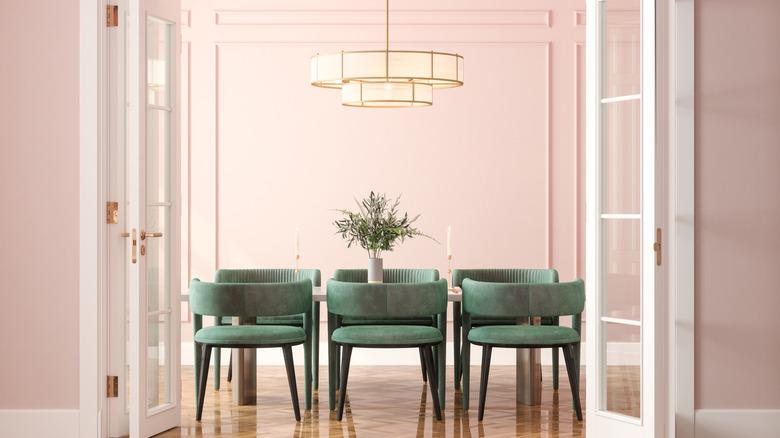 View through white french doors of a pink dining room with muted pink molded walls, modern tiered gold chandelier, and green velvet channelled dining chairs with black legs