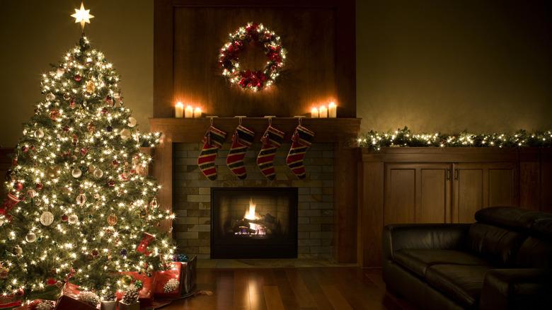 A living room decorated for Christmas. The only light from the room comes from the Christmas tree, candles on the mantle, lighted wreath, lighted garlands, and fire burning in the fireplace. Four stockings are hanging off the mantle.