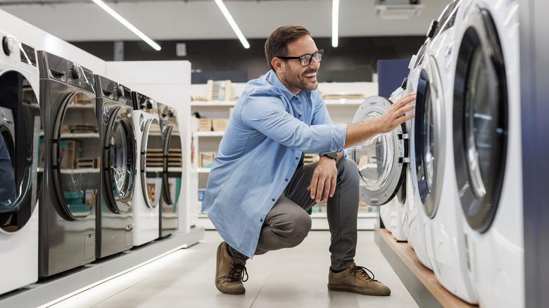 A smiling man is crouched in front of a new washing machine, touching the door. He is in the washing machine aisle of an appliance store.