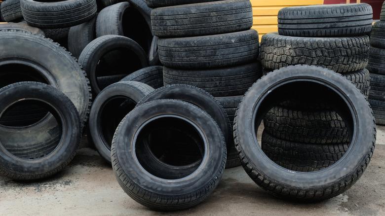 A collection of worn tires in various sizes are stacked outside, set on a concrete floor.