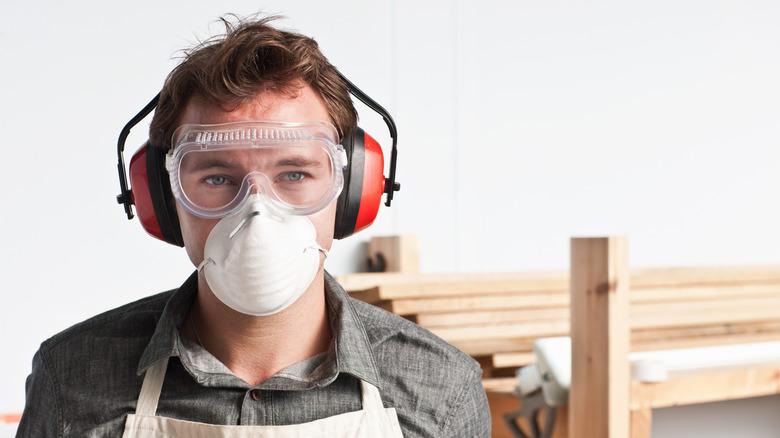 man wearing mask, goggles, ear protection with wood behind him