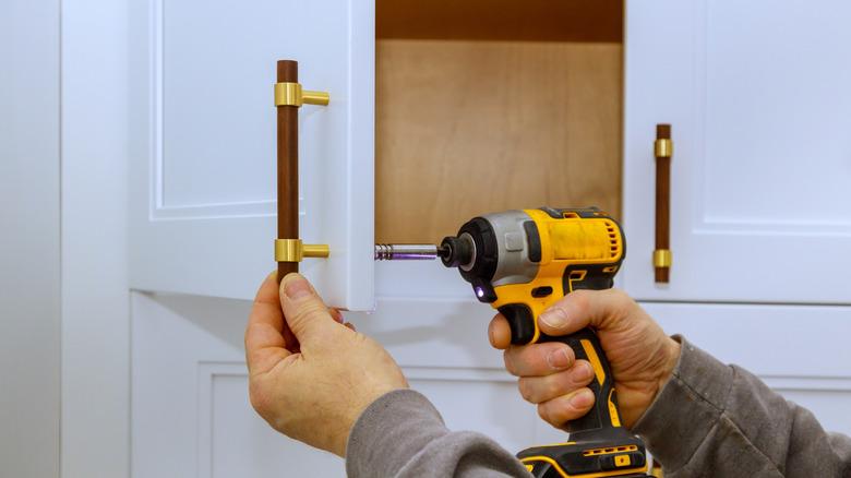 Person installing a wood and brass cabinet pull on an upper white kitchen cabinet door