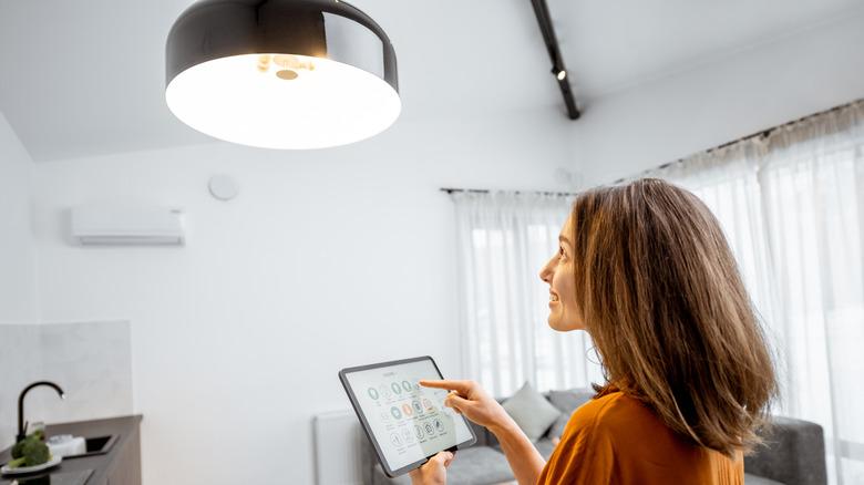 Over-the-shoulder of a woman holding her tablet and adjusting her smart lighting settings. she is looking upward toward an overhead light that's coming on in a white room with gray furniture.