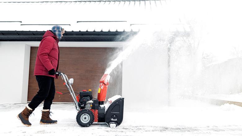 A medium shot of someone in a magenta jacket pushing a snow blower across their yard.