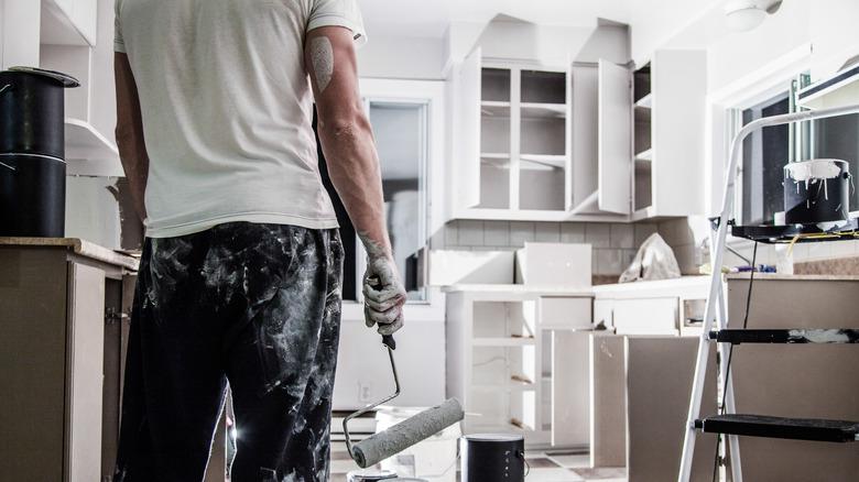 the back of a man holding a paint roller and covered in paint with an unfinished kitchen in the background