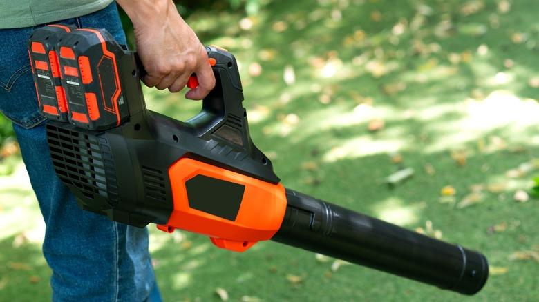 A man in jeans is shown from the waist-down, holding a black and orange leaf blower.