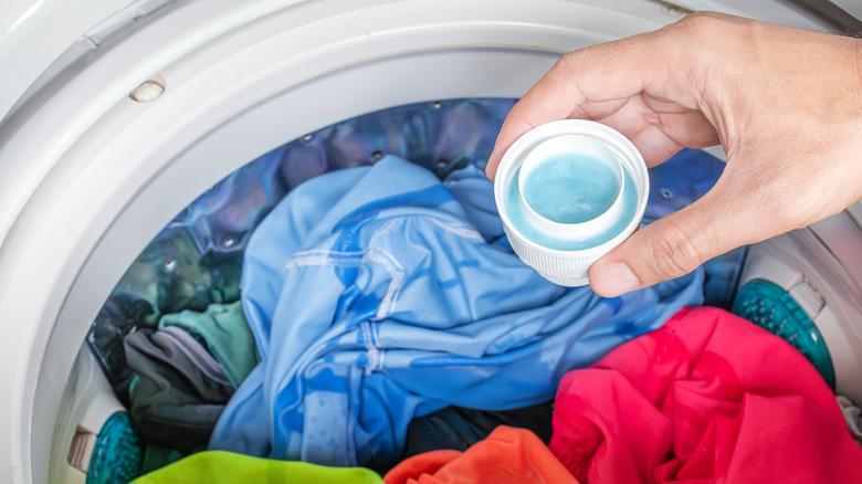 A hand about to pour blue laundry detergent into a washing machine filled with colorful clothes.