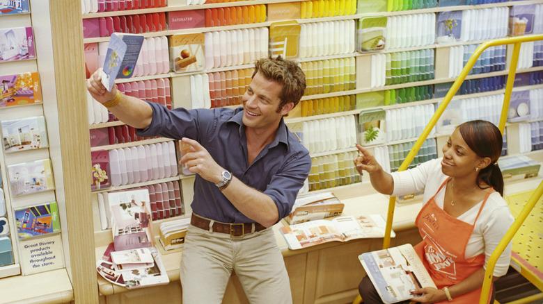 Nate Berkus holding up a paint sample in front of a wall of color swatches in an aisle of Home Depot