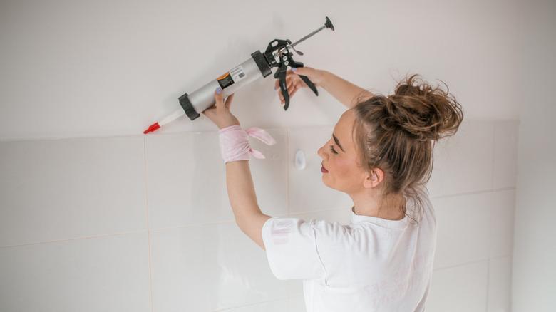 woman in a white shirt using a caulking gun to apply a line of caulk along the top of a white tile wall where it meets the drywall