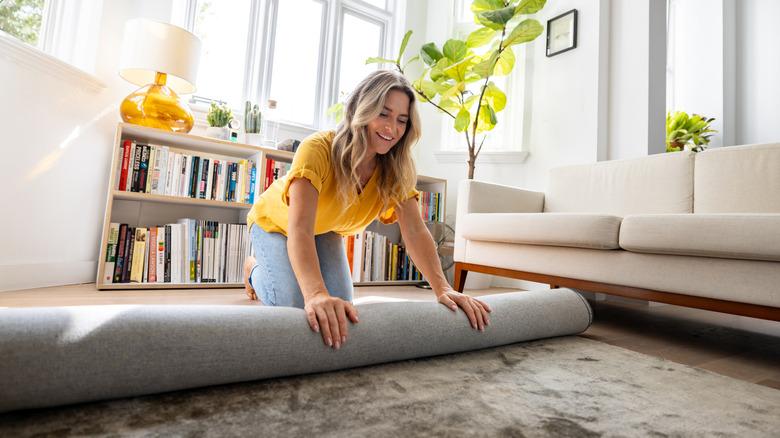 a woman with blonde hair, yellow shirt, and jeans rolls up a gray rug in a living room with a cream sofa and bookshelf under the windows