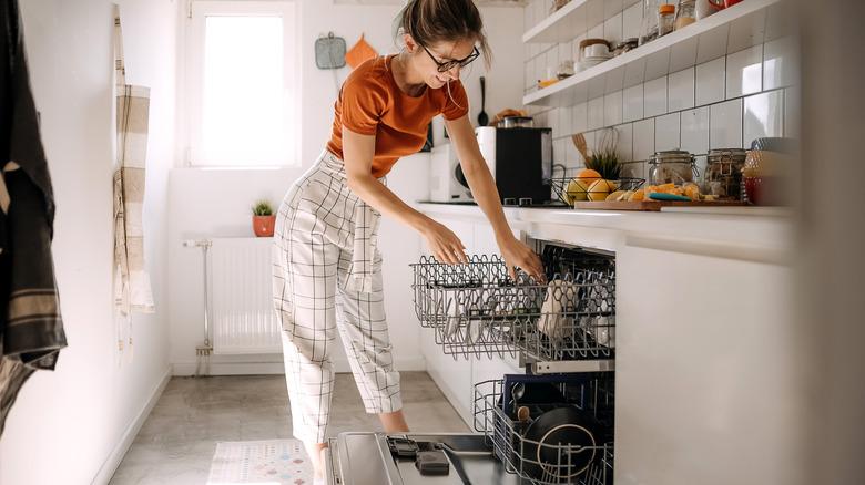 A woman with an orange shirt and capris is loading dishes into a dishwasher in a white kitchen.