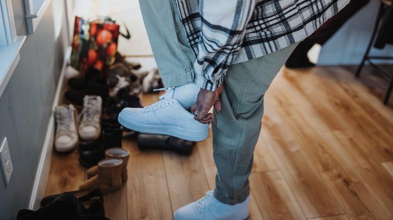 man putting on a pair of shoes in an entryway with wood floors and a pile of other shoes on the floor behind him