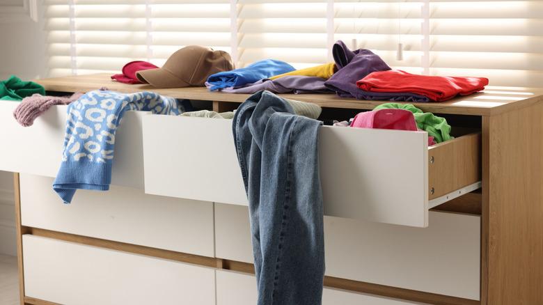 lowboy dresser with wood surround and white drawer fronts has the top drawers open and overflowing with clothing clutter