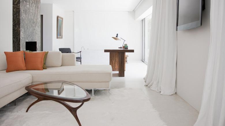 living room with white floor to ceiling drapery next to a glass sliding door, cream sectional with orange pillows, white fur rug, and white floors