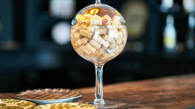 A tall glass decor piece, filled with corks, sitting on the table.