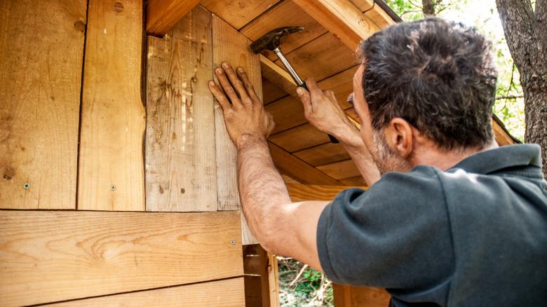 man building treehouse, placing hand on wood while using hammer