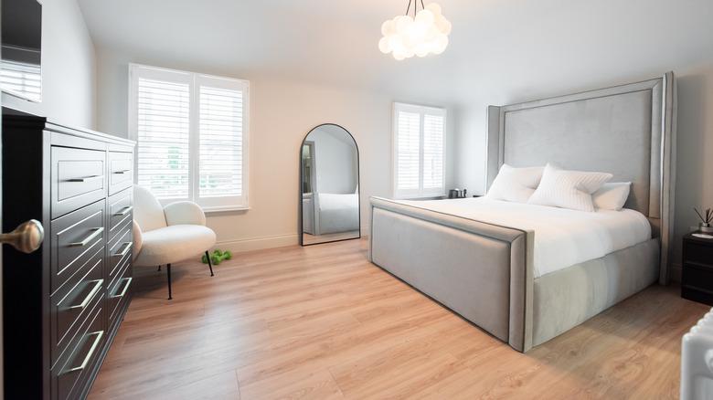 A light-filled bedroom with a neutral color scheme. The room interior consists of a bubble chandelier, light wood floors, a gray upholstered bed with white linens, a floor mirror, white chair, and dark wood dresser.