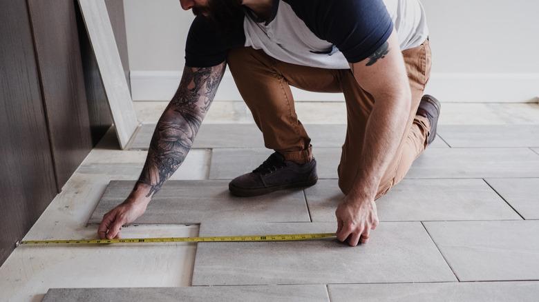 A man with a beard and tattoo-sleeve is measuring the distance to the wall while installing tile flooring.