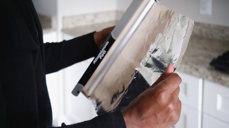 A pair of hands pulling out a large piece of aluminum foil from a roll in a kitchen.
