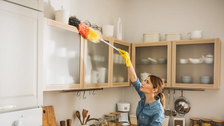woman in jean shirt dusting the top of a cabinet in a beige kitchen