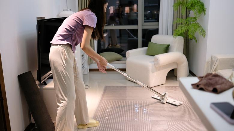 Woman using mop in a living area with TV and armchair.