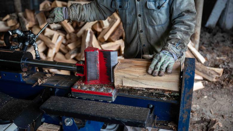 A red and blue log splitter is being operated by someone in a denim jacket and gloves.