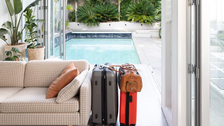 Looking out the back of a living room filled with tropical plants, a couch, and some suitcases, indicating it is a vacation home. The glass doors are open onto a patio with a pool.