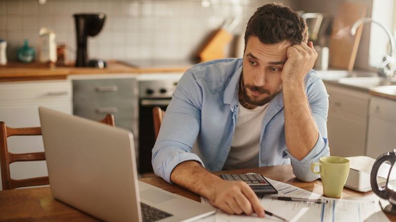 A stressed-looking man is sitting at his kitchen table looking at a laptop. A cup of coffee, bills, and a calculator are spread across the table.